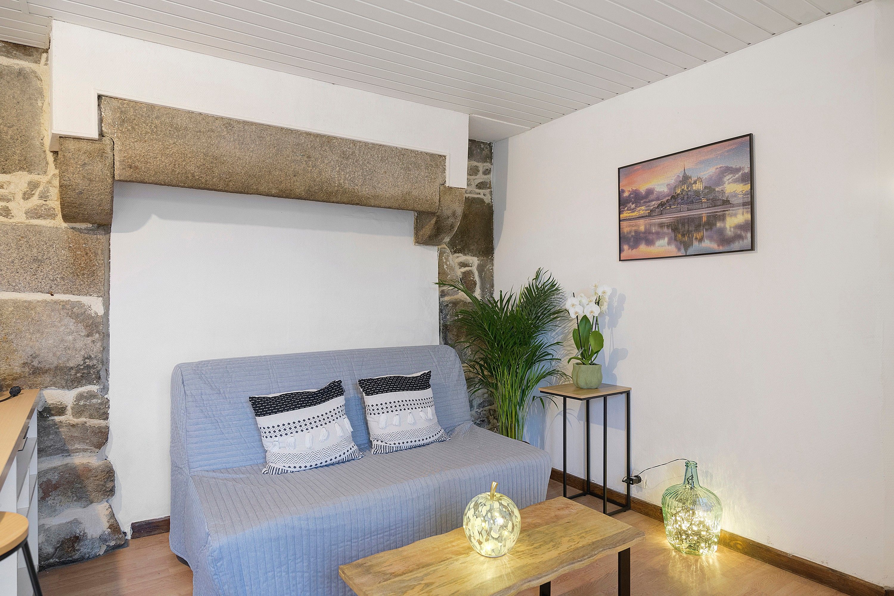 A room with a stone wall, a blue sofa with patterned cushions, a wooden table with a glass vase, and a painting on the wall.