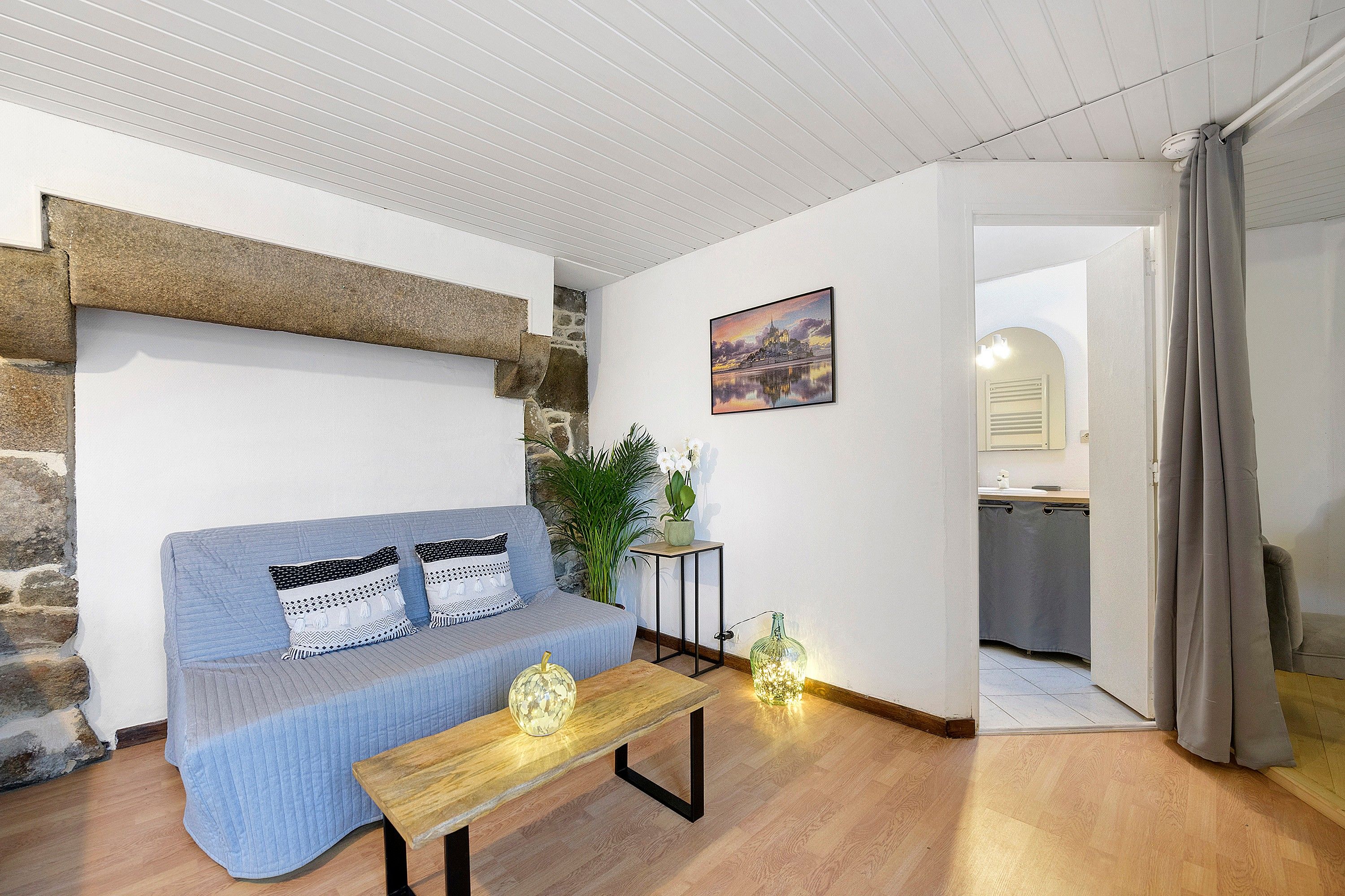 A cozy living room with a blue sofa, a wooden coffee table, a stone wall, a painting, and a view into the kitchen.