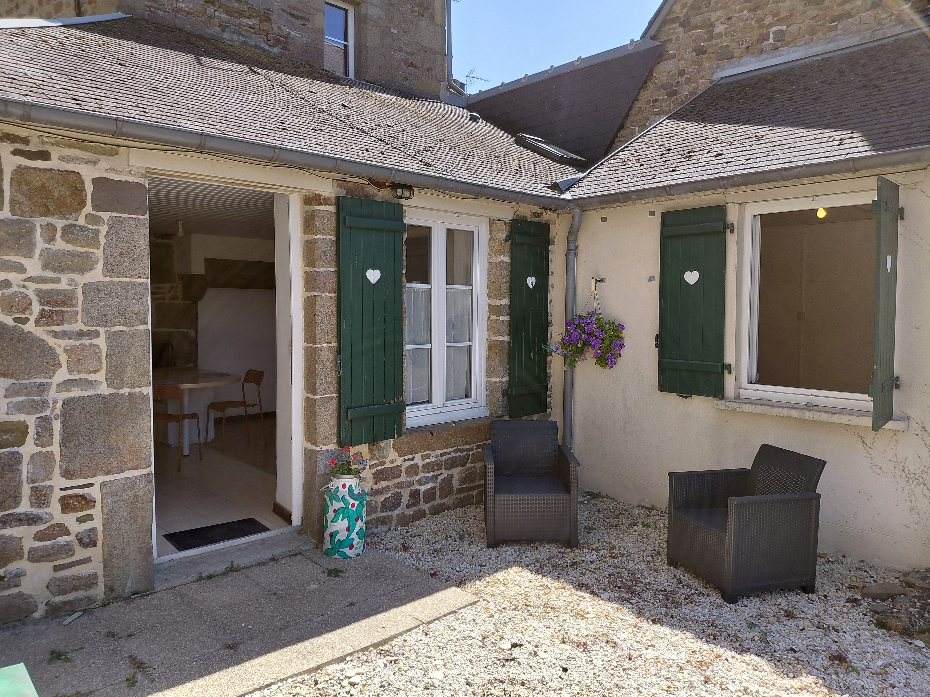 Un bâtiment en pierre avec un toit en tuiles, un petit patio avec deux chaises en osier et une porte donnant sur un espace intérieur.