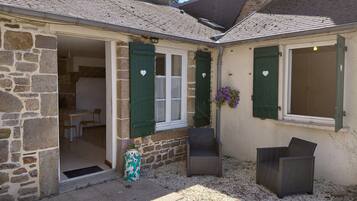 Un bâtiment en pierre avec un toit en tuiles, un petit patio avec deux chaises en osier et une porte donnant sur un espace intérieur.
