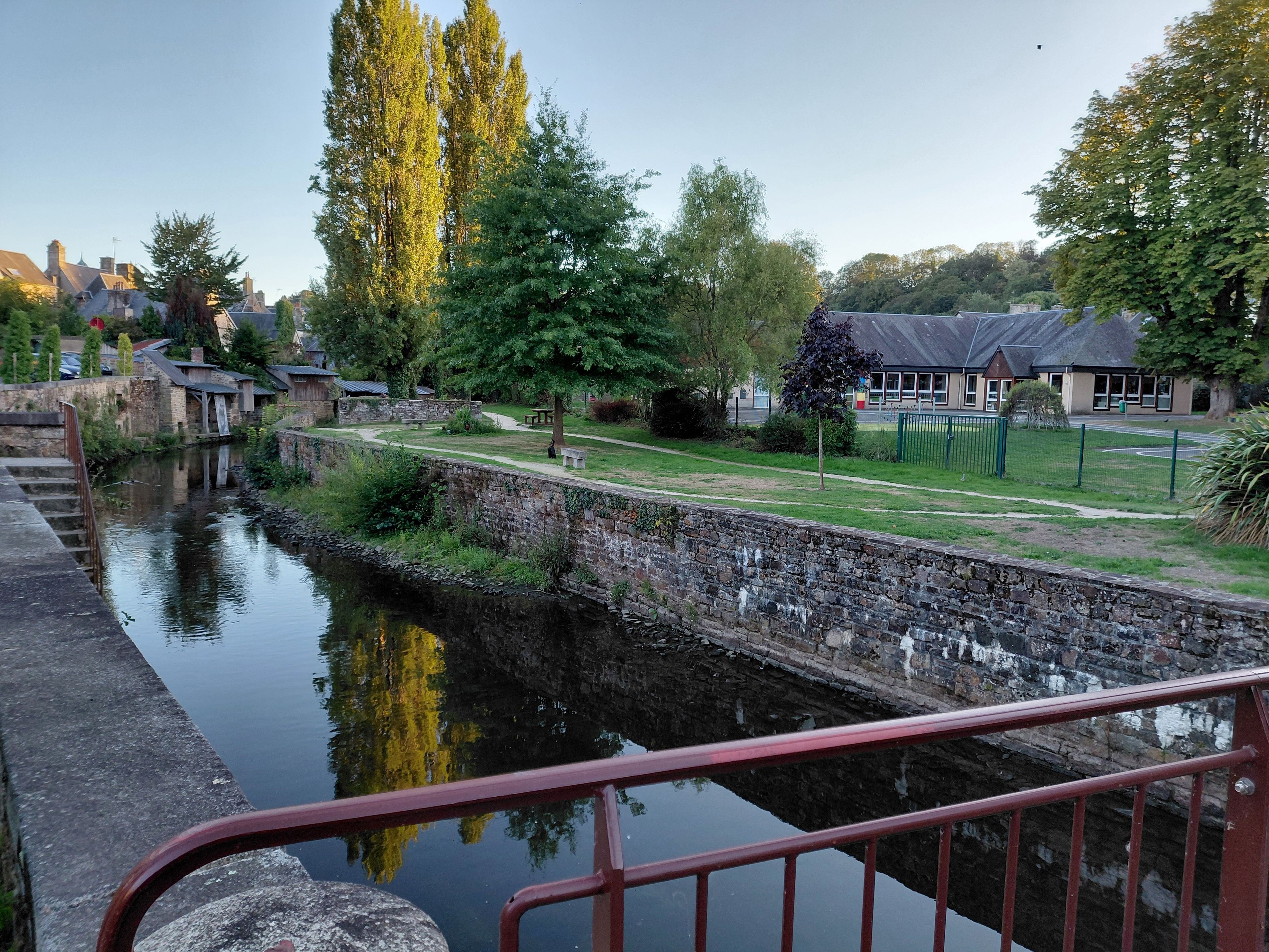 Une rivière paisible, avec un mur de pierres et une rambarde rouge au premier plan.