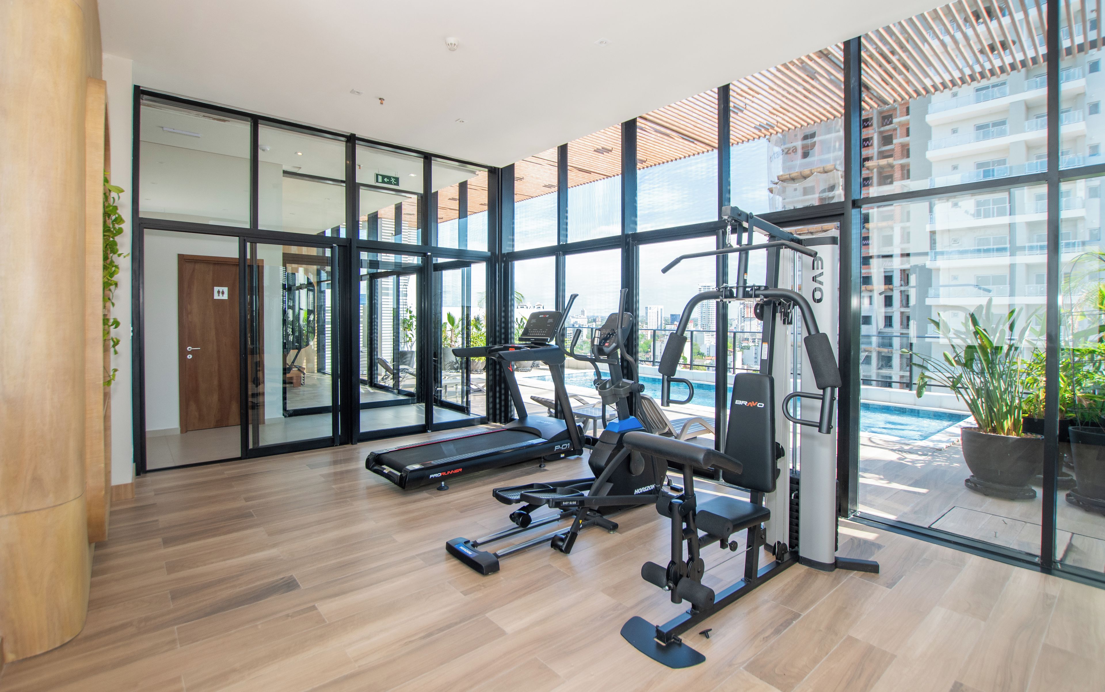 Un gimnasio moderno con equipo de ejercicio, ventanas amplias y vistas a un paisaje urbano.