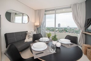 A modern living room with a sofa, dining table set for two, a potted plant, and a view of the cityscape through large windows. - Corporate Stays Habitalis Mburucuya- 15E (Asunción)