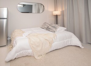 A neatly made bed with a white comforter and two pillows, a bedside lamp, a mirror, and a modern refrigerator. - Corporate Stays Habitalis Mburucuya- 15E (Asunción)