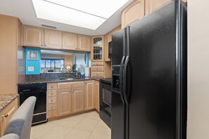 A kitchen with a black refrigerator, wooden cabinets, a blue countertop, and a skylight. - Carlos Pointe #224 | Beautiful Beachfront Condo, Gulf Views + Pool 🌊 (Fort Myers Beach)