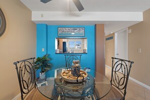 A dining area with a glass table surrounded by metal chairs, a decorative centerpiece, and a blue accent wall. - Carlos Pointe #224 | Beautiful Beachfront Condo, Gulf Views + Pool 🌊 (Fort Myers Beach)