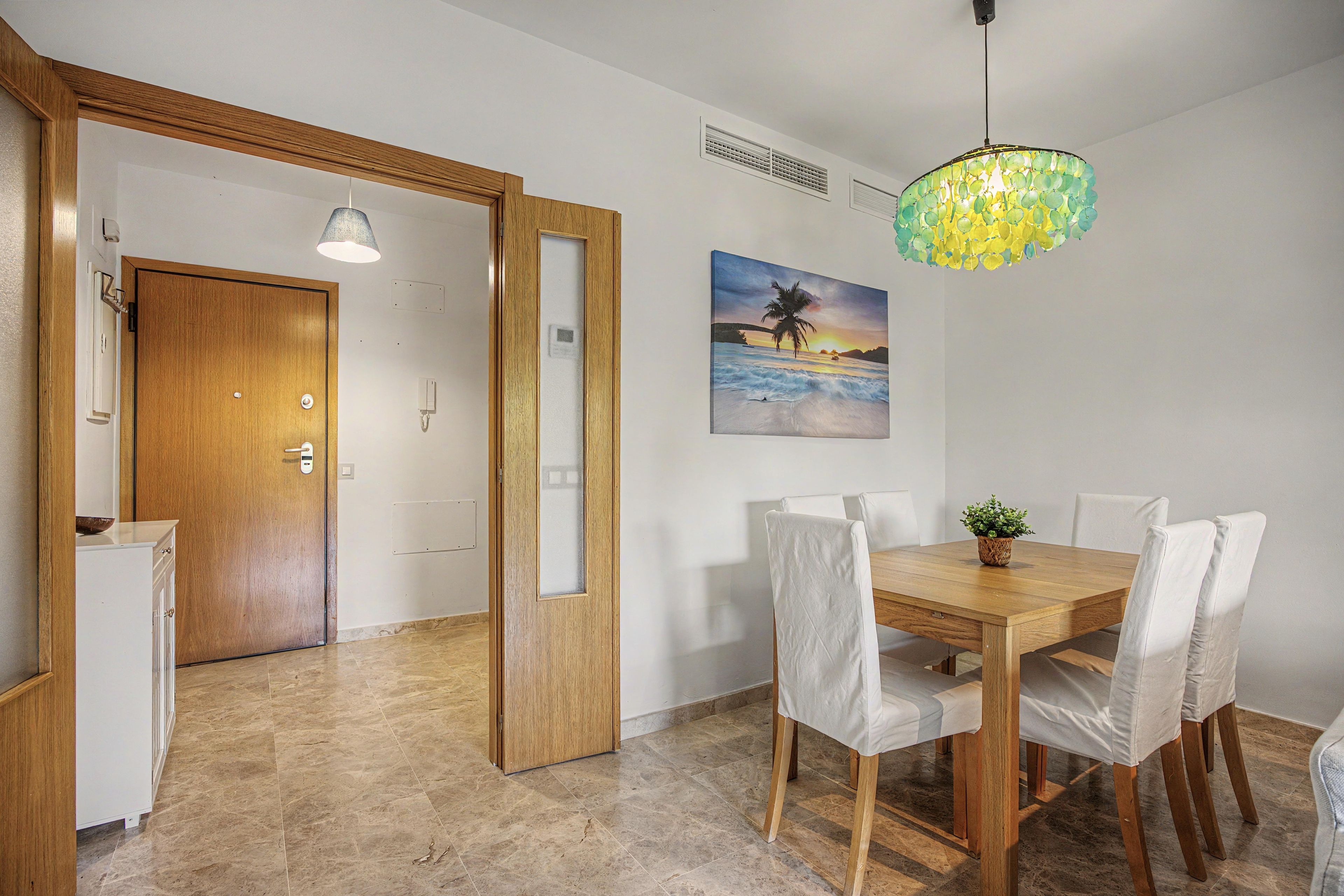 A dining area with a wooden table and chairs, a painting of a beach scene, and a ceiling light fixture.