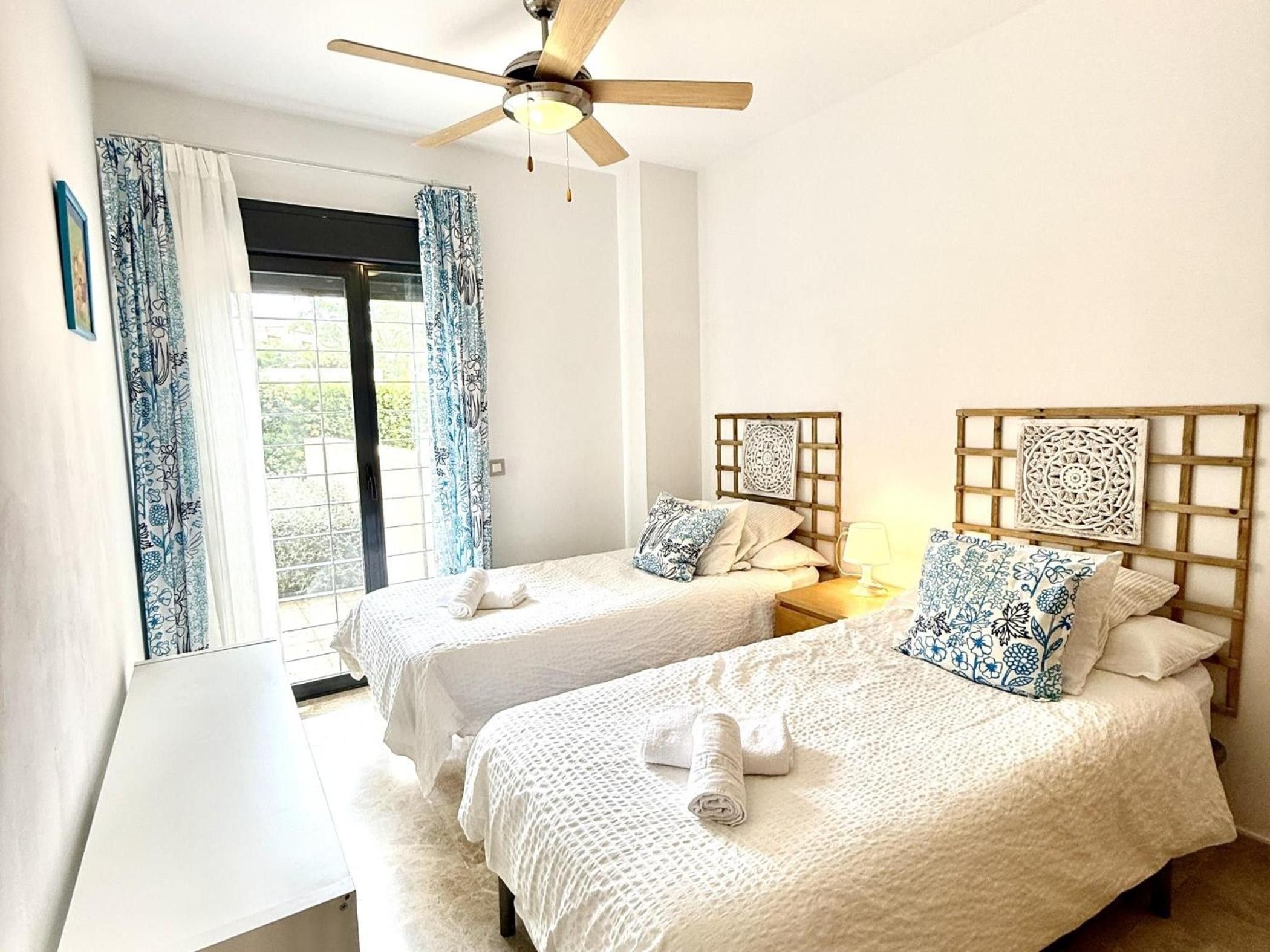 Habitación de hotel con dos camas, ventilador de techo y ventana con cortinas.