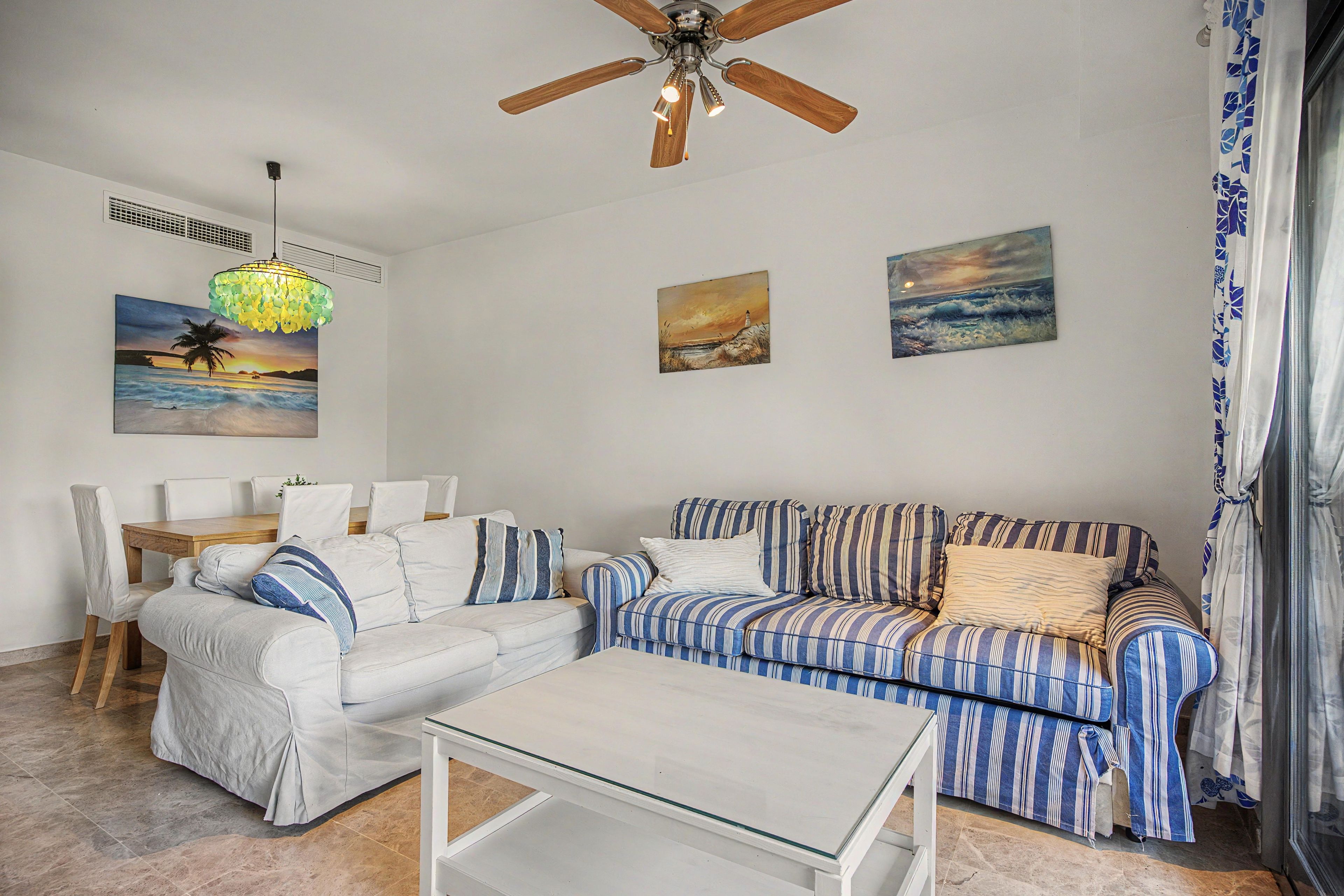 A living room with a ceiling fan, a dining table with chairs, a sofa, a striped couch, and a glass coffee table.