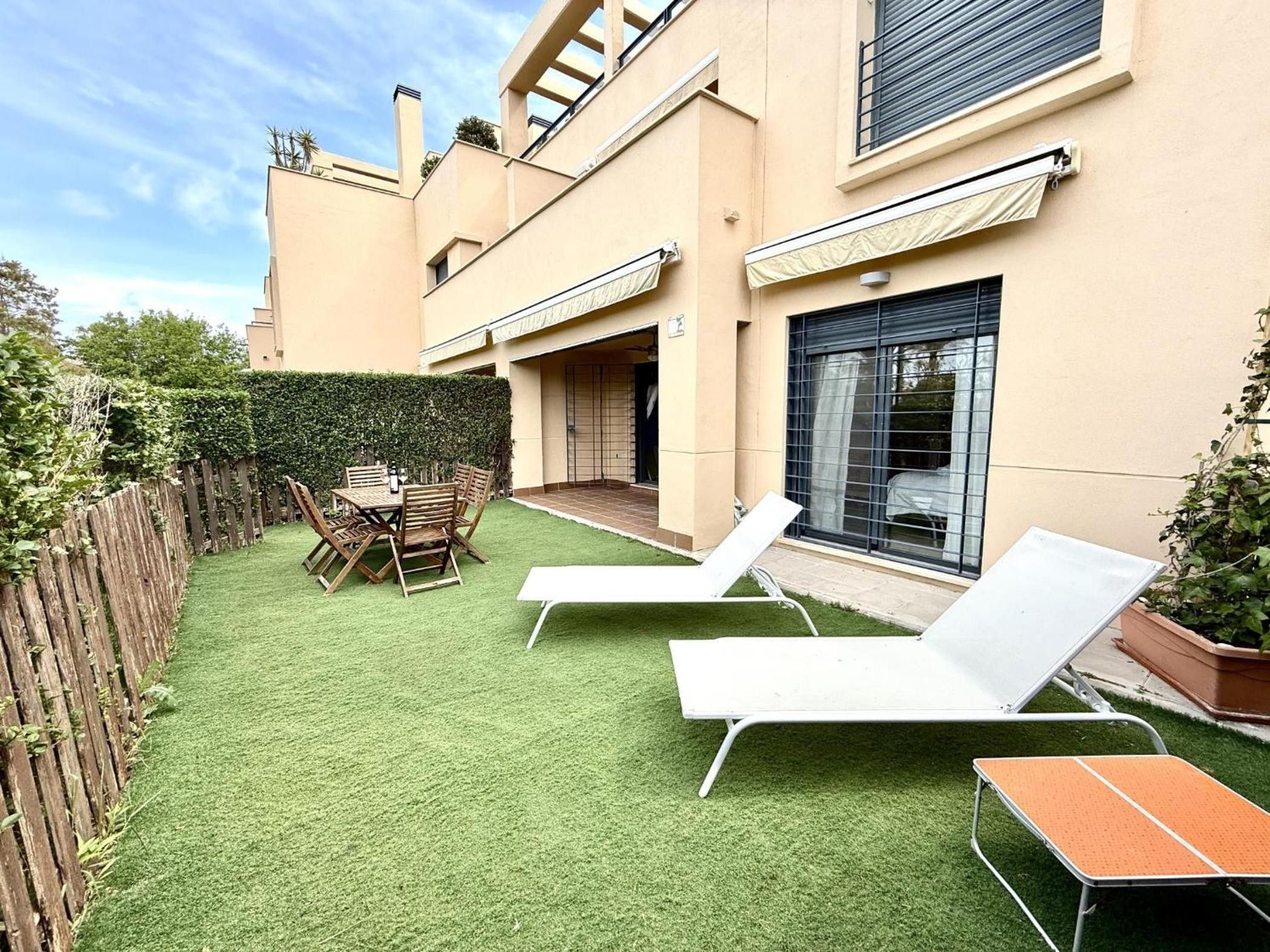 Un patio trasero con césped artificial, una mesa de madera, dos sillones de descanso y una valla de madera.