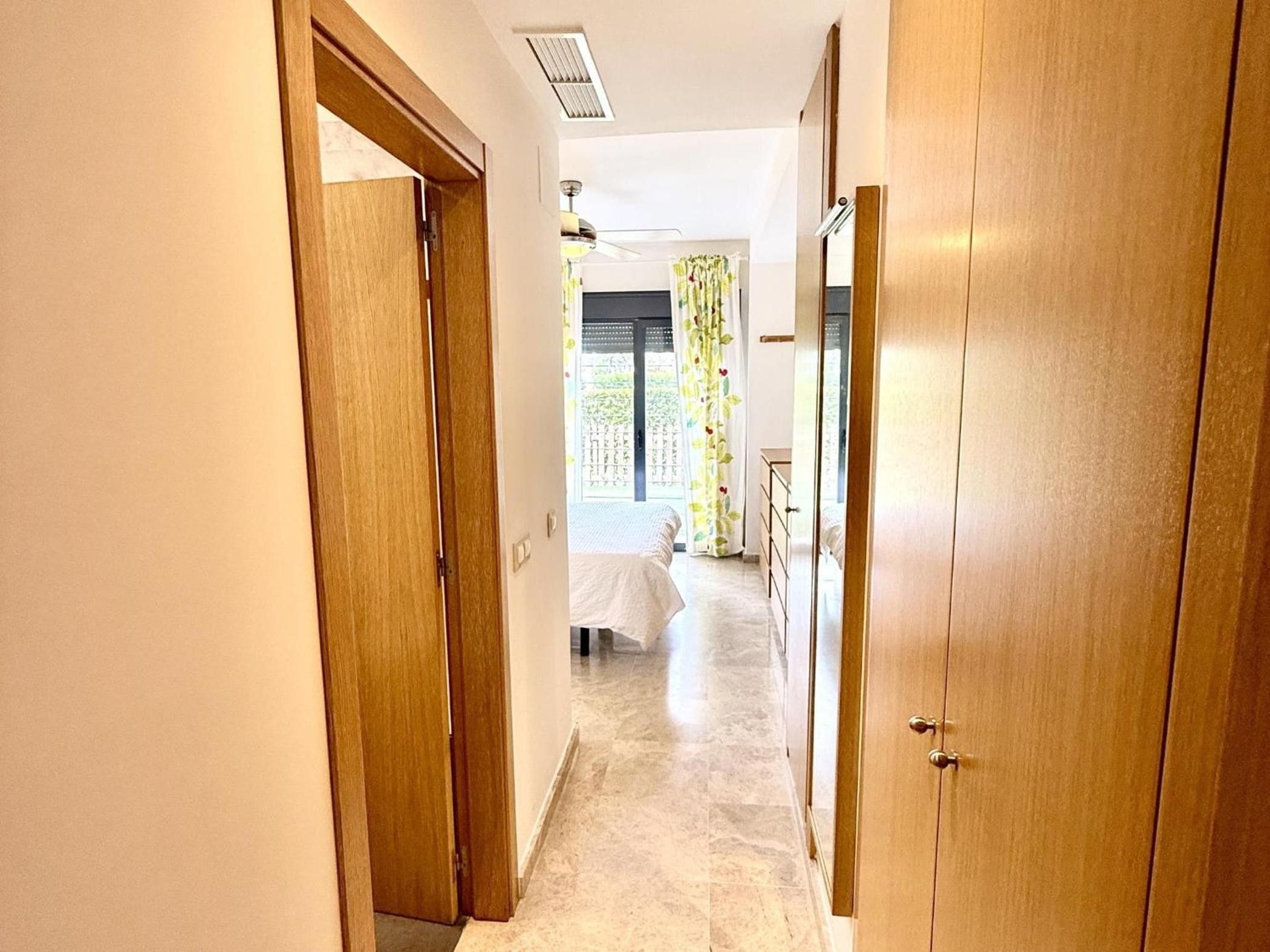 Un pasillo con puertas de madera, una cama con cubrecama blanco y una ventana con cortinas florales.