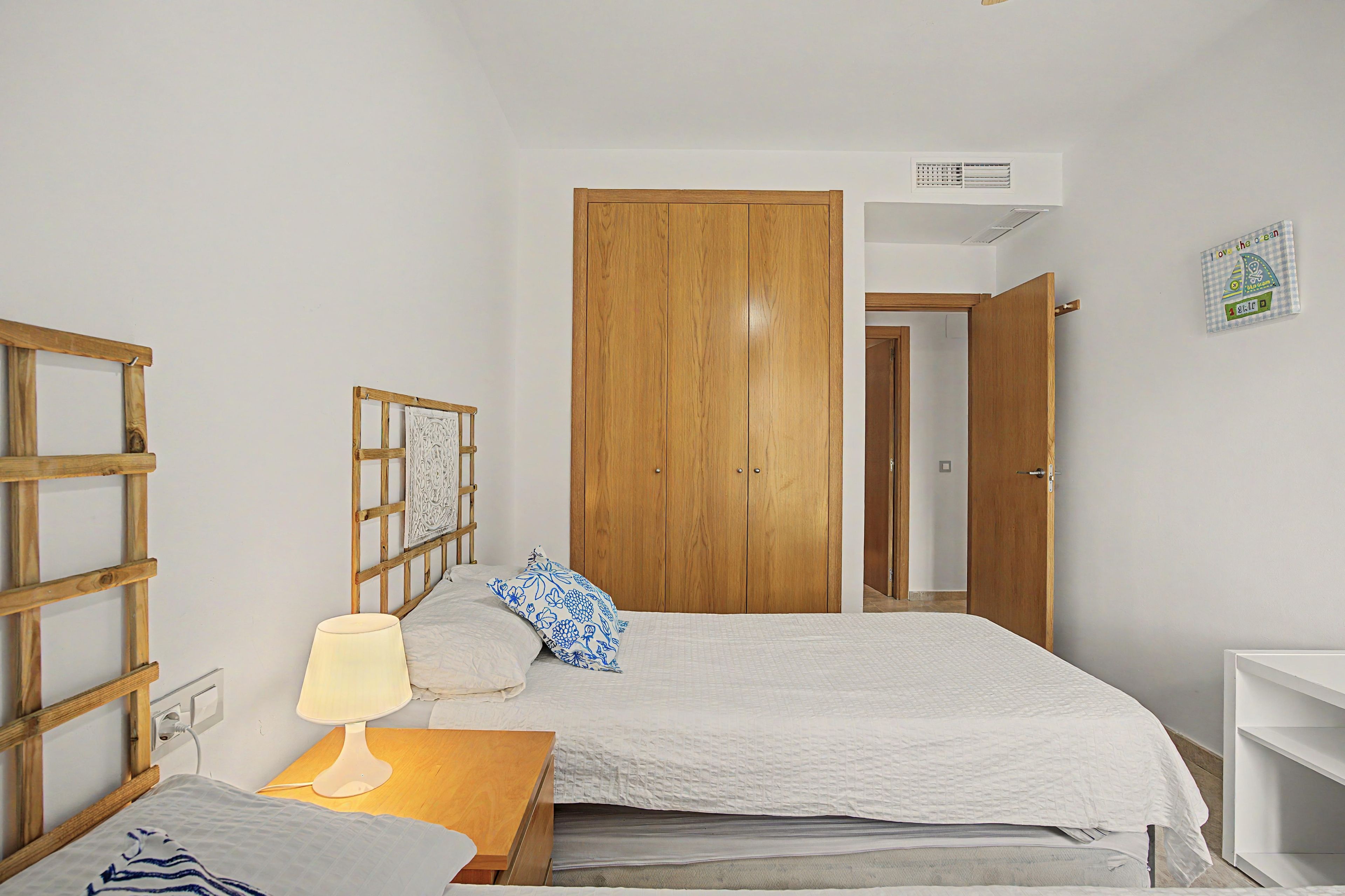 A bedroom with a wooden wardrobe, a bed with a white duvet, a bedside table with a lamp, and a wall-mounted air conditioning unit.
