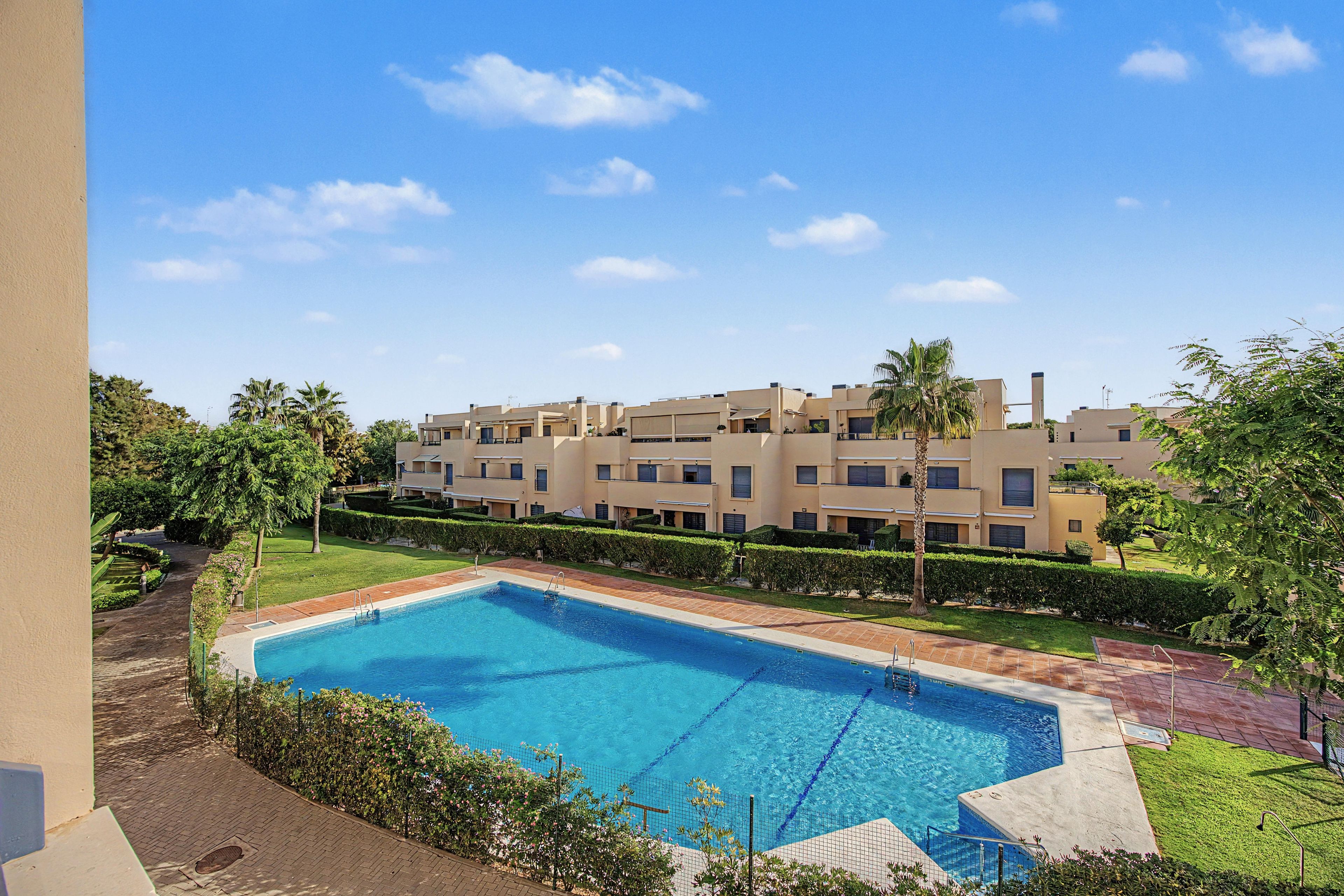 A swimming pool with a clear blue water surface, surrounded by a well-maintained lawn and a row of buildings in the background.