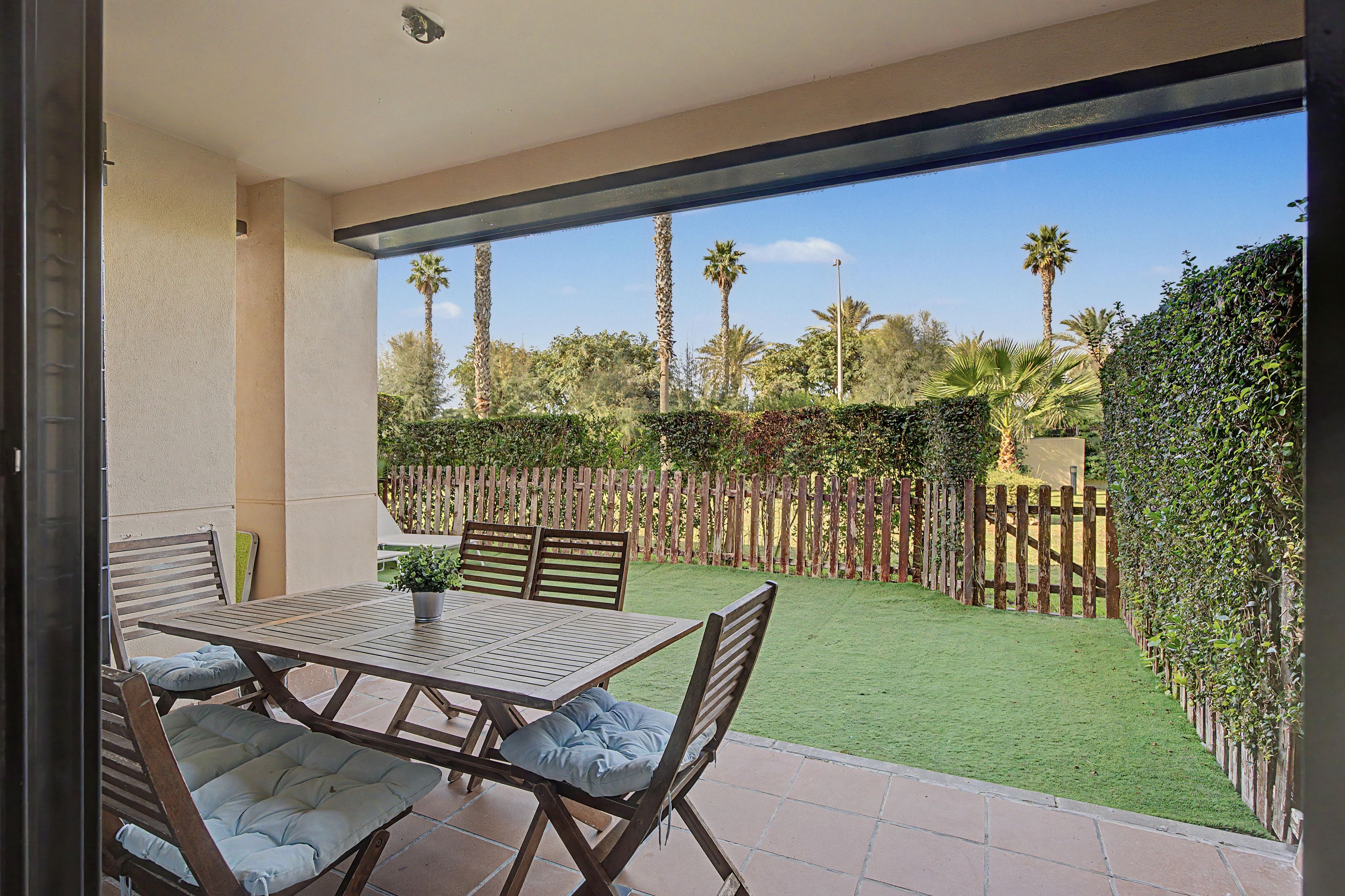 An outdoor patio with a table and chairs, a view of a well-maintained garden with a wooden fence, and a clear sky.