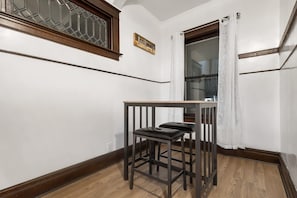 Una stanza d'angolo con pavimento in legno, un tavolino con una sedia, una finestra con tende e un cartello di legno appeso al muro.