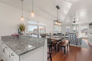A spacious kitchen with a granite countertop, dining area with wooden chairs, and a view of the outside through large windows. - Sea Dunes D1: Coastal Retreat with Ocean Views (Kitty Hawk)