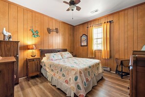 Une chambre avec des lambris en bois, un lit recouvert d’une courtepointe à motifs floraux, une table de chevet avec une lampe et un ventilateur de plafond.