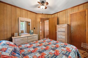 A bedroom with wooden panel walls, a bed with a patterned quilt, a dresser, a mirror, and a ceiling fan. - Sea Bird: Live Oak Tree Swing (Kill Devil Hills)