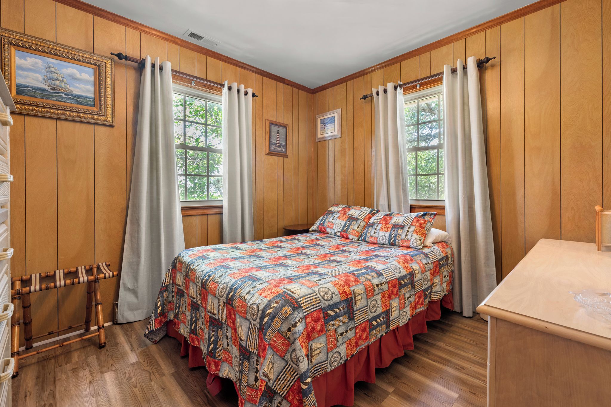 Een slaapkamer met een houten wandpaneel, een bed met een kleurrijke quilt, twee ramen met gordijnen en een klein tafeltje.