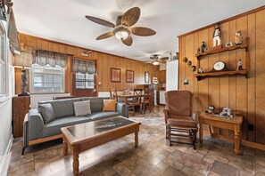 Ein Wohnzimmer mit einer Couch, einem Couchtisch und einem Holzstuhl. Es gibt einen Essbereich mit Tisch und Stühlen. Die Wände sind mit Holz verkleidet, und es gibt Regale mit Figuren und einer Uhr.