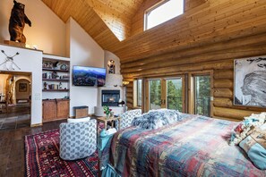 A log cabin interior with a fireplace, a television, a large bed, and a view of the outdoors through the windows. - Sundance Lodge in Angel Fire (Angel Fire)