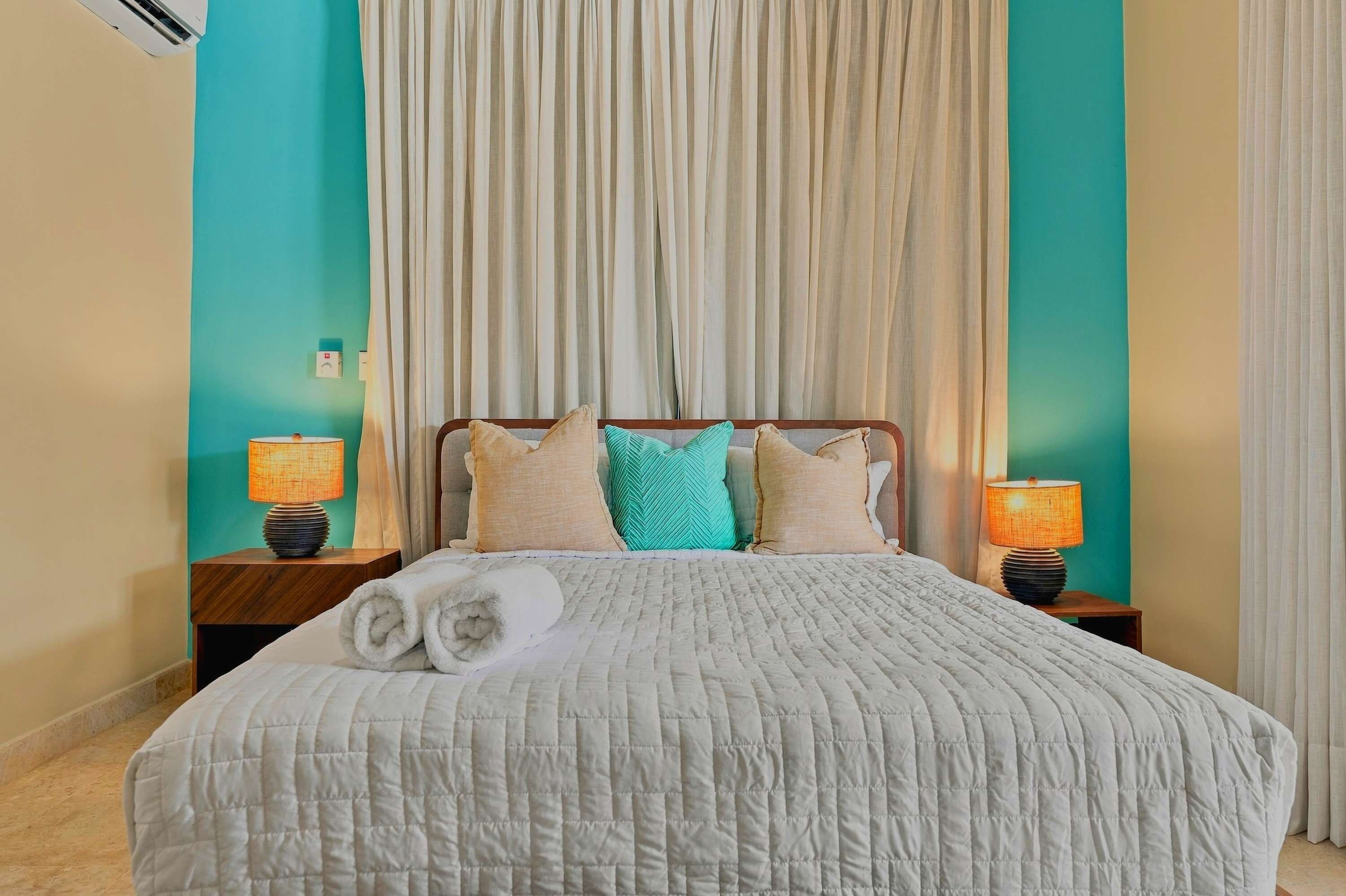 A neatly made bed with a white quilt, two bedside lamps, and a set of towels.