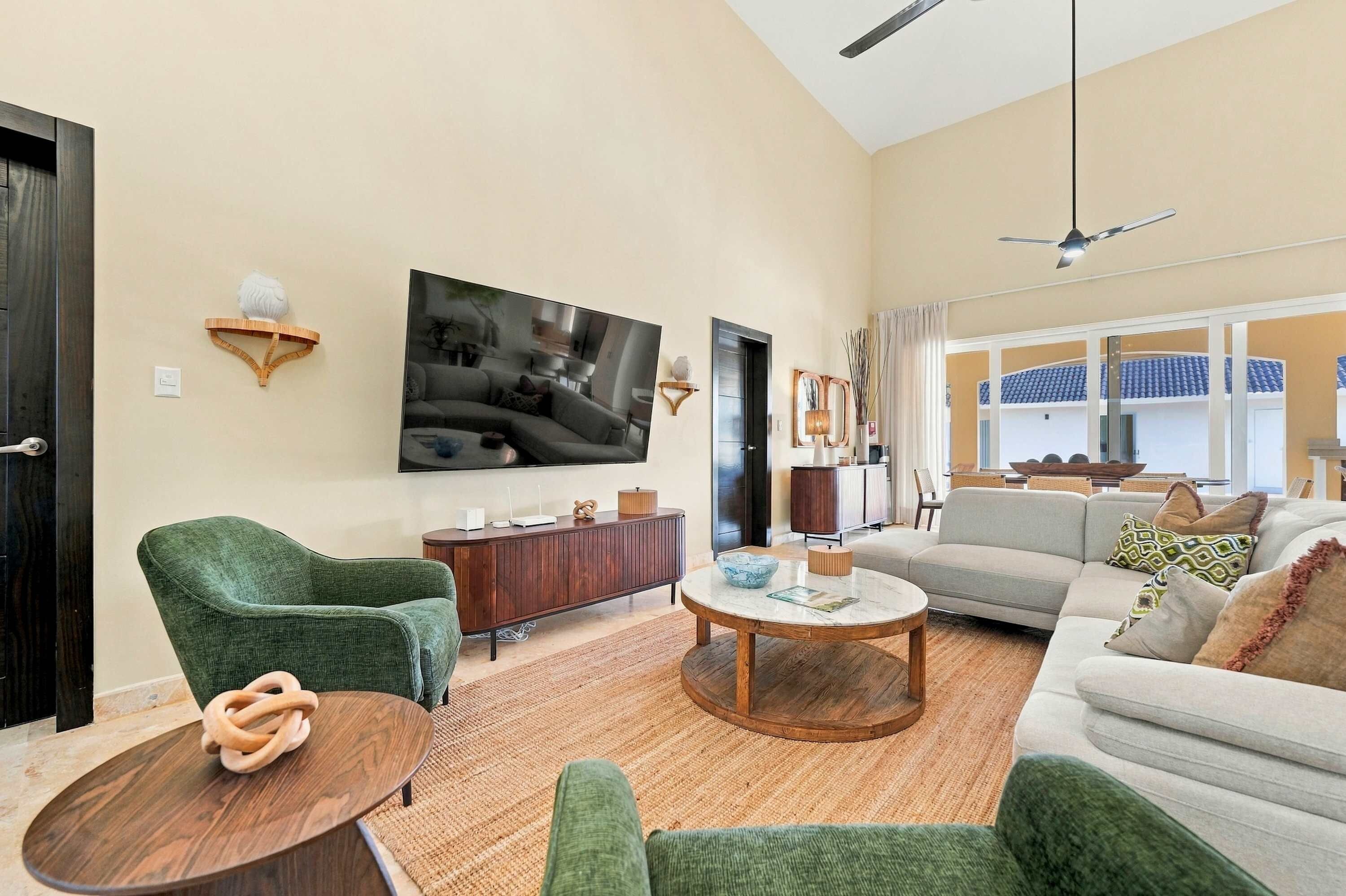 A modern living room with a flat-screen TV, green armchairs, a round coffee table, and a sofa with patterned cushions.