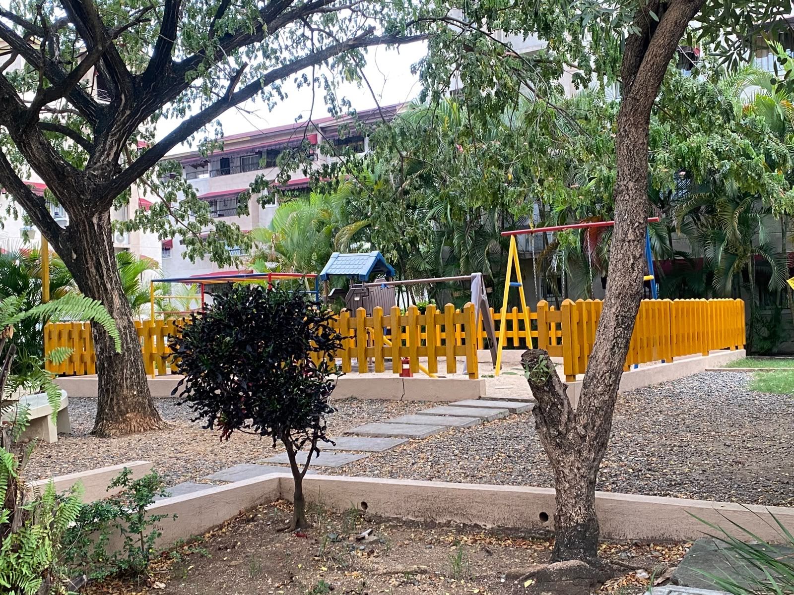 A playground with yellow fencing, a small tree, and a building with a red roof in the background.