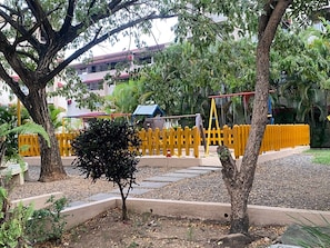 A playground with yellow fencing, a small tree, and a building with a red roof in the background.