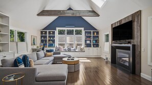 A modern living room with a sectional sofa, a round coffee table, and a fireplace. There are built-in shelves with decorative items and a large window providing natural light. - Private East Hampton Oasis with Heated Pool & hot tub, Modern Interiors,... (East Hampton)