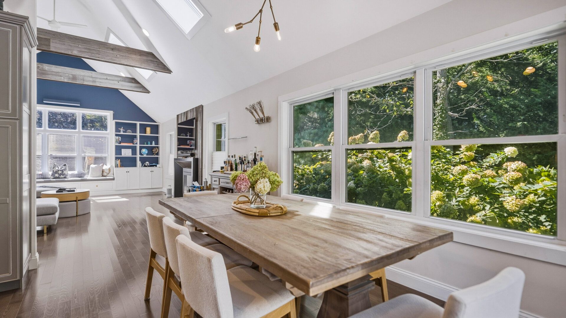 Un vaste espace dédié à la salle à manger, avec une grande table en bois, des chaises blanches et une vue sur une végétation luxuriante grâce à de grandes fenêtres.