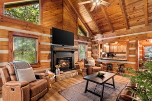 A log cabin interior with a fireplace, a television, a sofa, and a kitchen area. - Updated Cabin • 3 Queens • Fire Pit • 0.2 Mi to Smoky Mountain Park • Bike trail (Townsend)