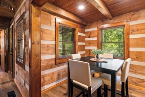 A log cabin interior with a dining area featuring a wooden table and chairs, large windows with blinds, and a ceiling with exposed beams. - Updated Cabin • 3 Queens • Fire Pit • 0.2 Mi to Smoky Mountain Park • Bike trail (Townsend)