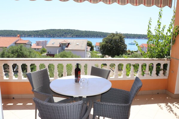 Ein runder Tisch mit Korbstühlen auf einem Balkon mit Blick auf einen See, im Hintergrund Boote und Häuser.