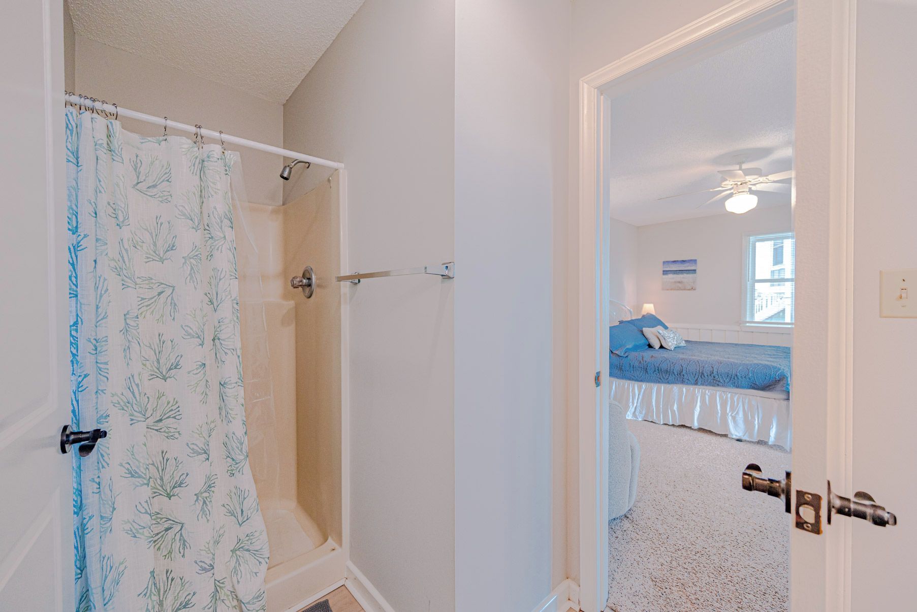 A bathroom with a shower, a door leading to a bedroom with a bed and a window.