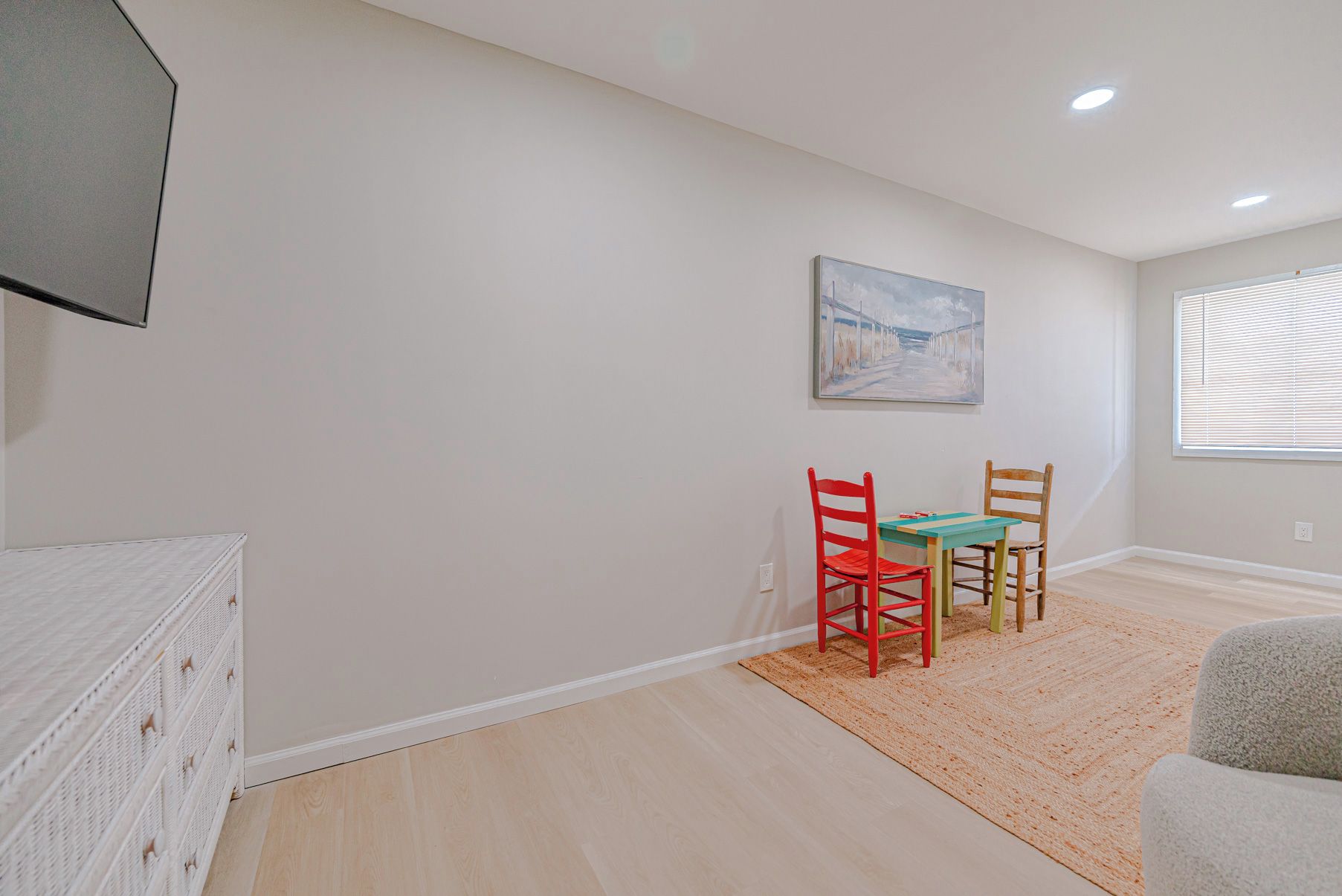 A room with a bed, a table with chairs, a painting on the wall, and a window with blinds.