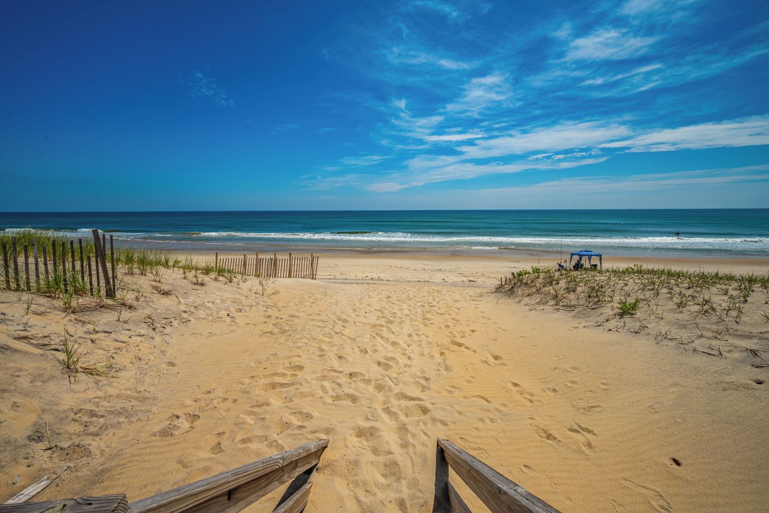 A beach with a wooden walkway leading to the water, a fence on the left, and a small structure on the right.