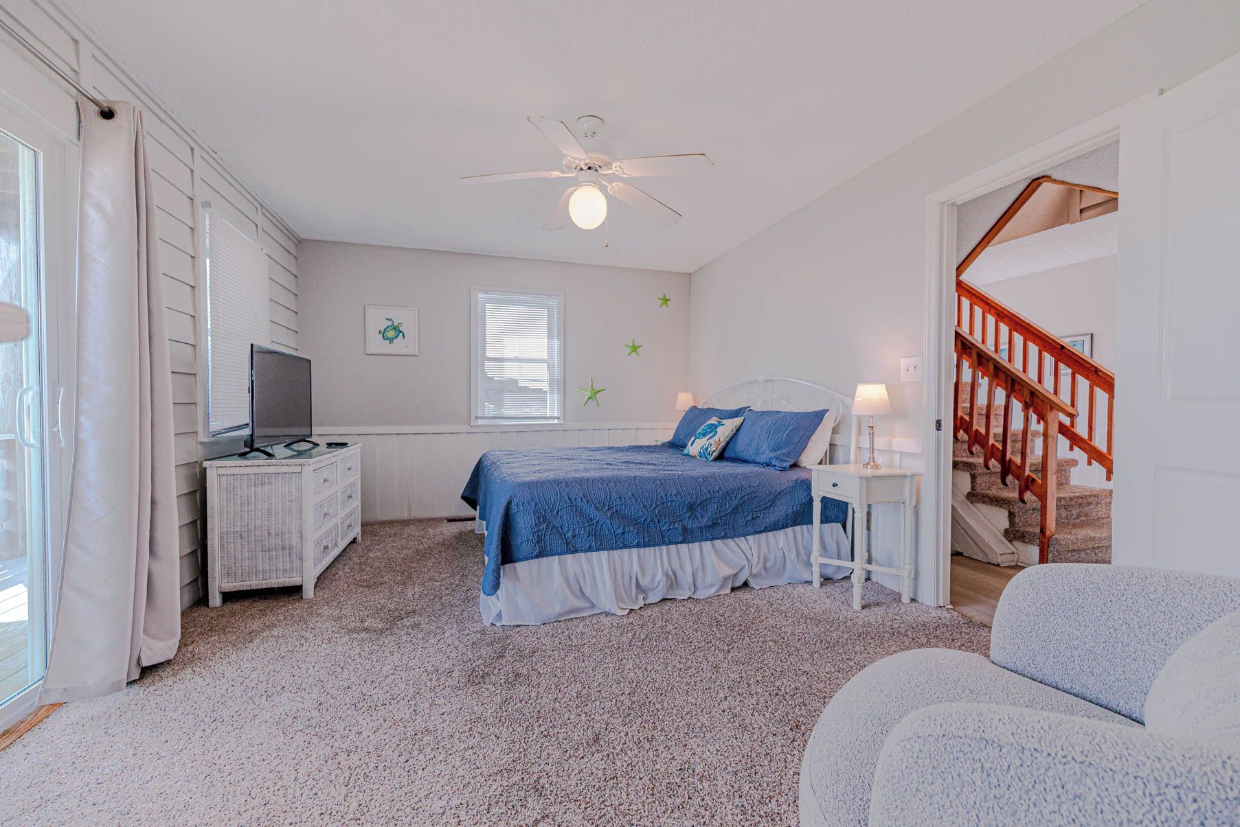 A bedroom with a bed, a desk, a television, a ceiling fan, a staircase, and a window with blinds.