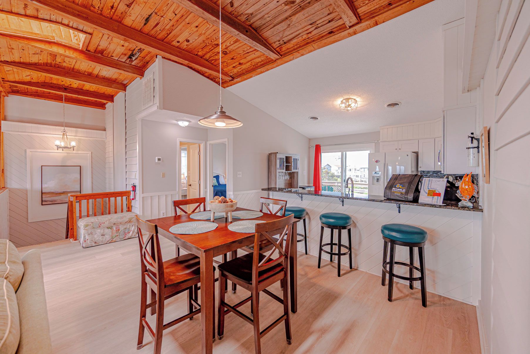 A spacious kitchen with a wooden ceiling, a dining area with a wooden table and chairs, and a view of the outside through a window.