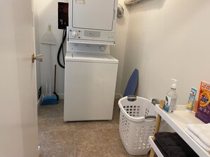 Zona de lavandería con lavadora, escoba, cesto de ropa y productos de limpieza.