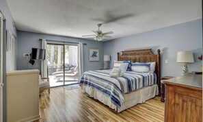 Un dormitorio con cama, ventilador de techo, televisor y vista a través de un ventanal.