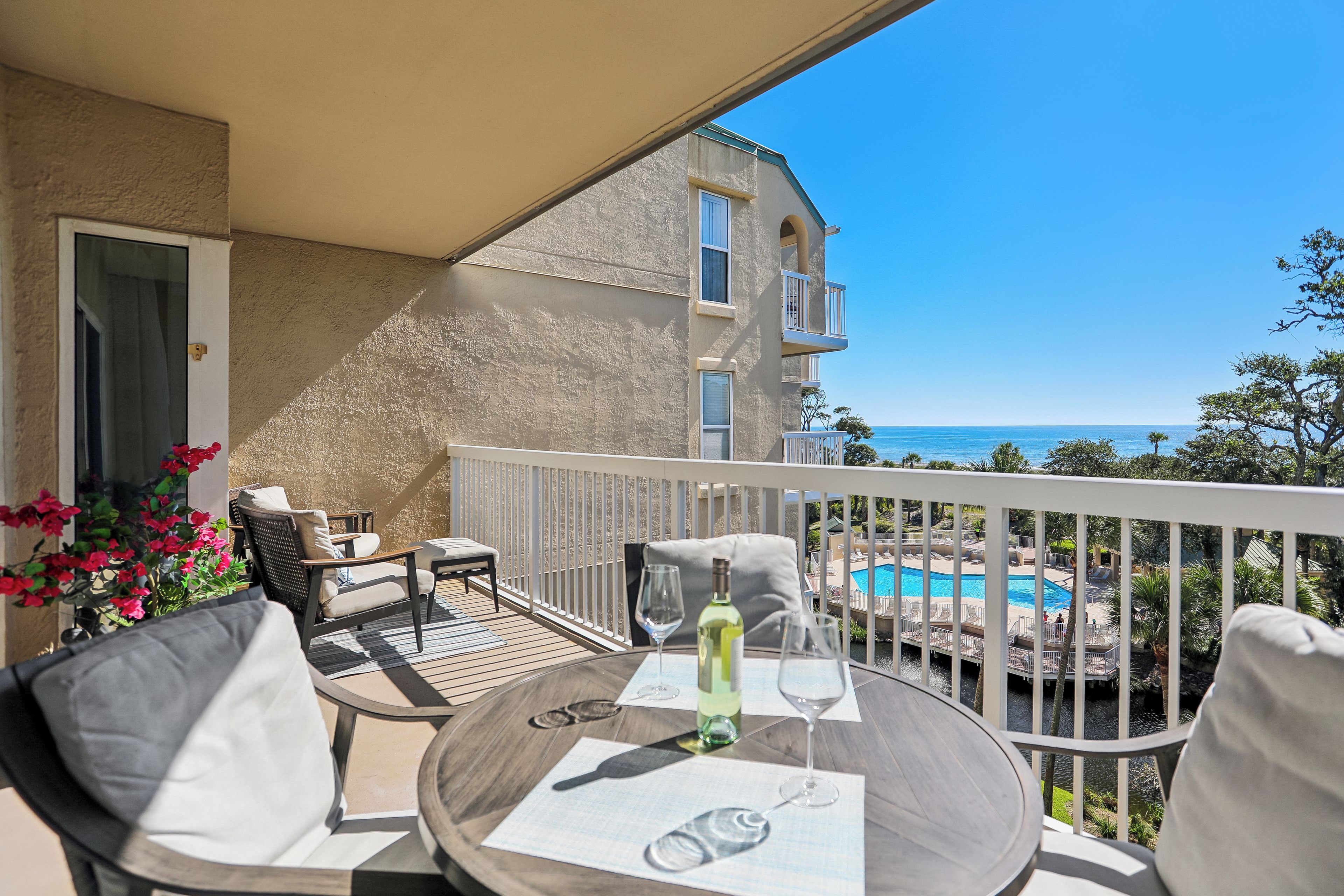 A balcony with a table set for two, a bottle of wine, and a glass. The balcony overlooks a swimming pool and a building with a view of the sea.