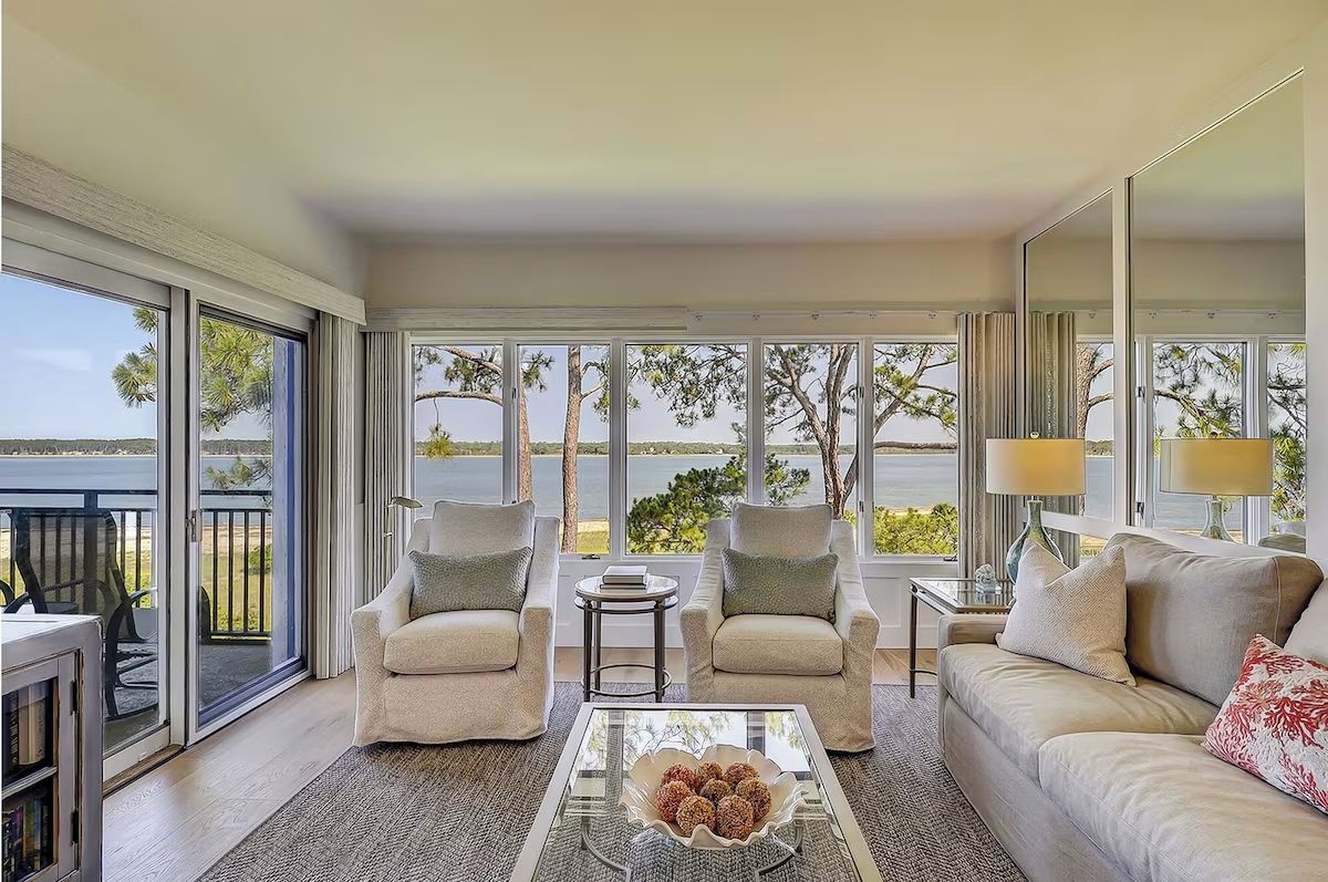 A spacious living room with a large window, a sofa, two armchairs, a coffee table, and a side table with a lamp.