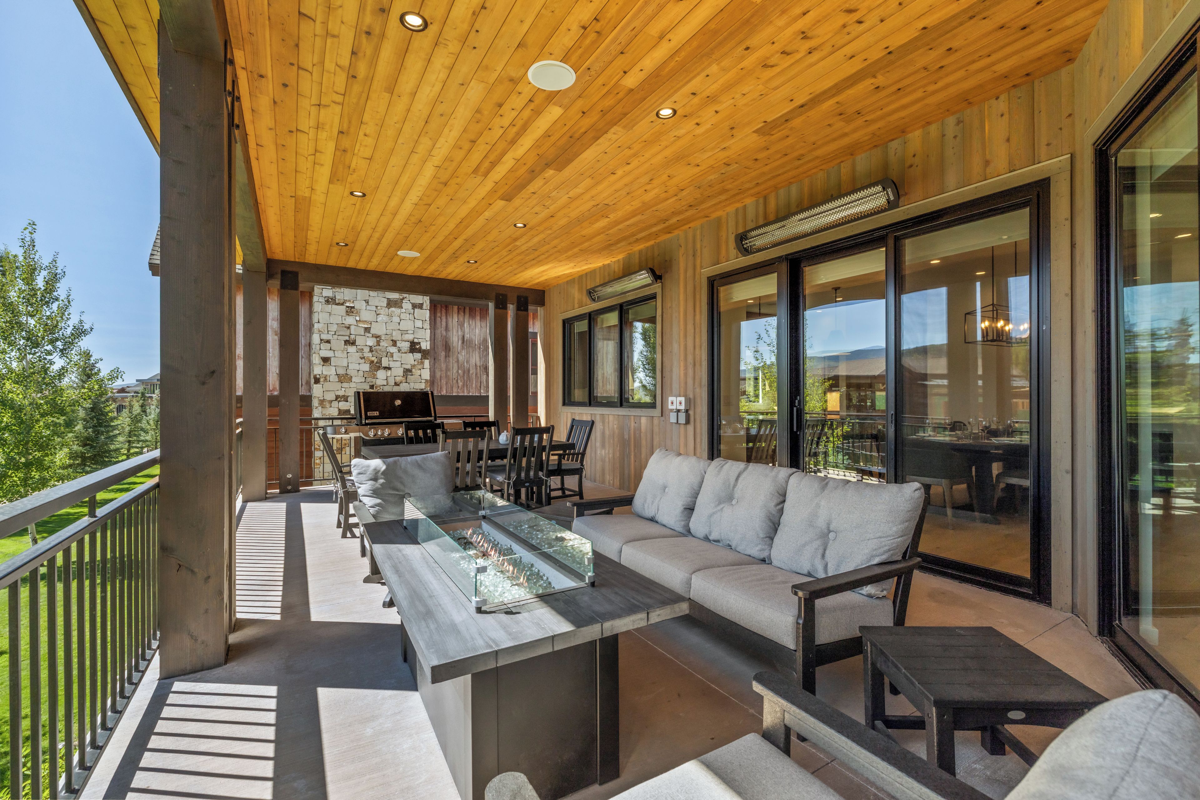 An outdoor patio with a wooden ceiling, a stone wall, a barbecue grill, and a seating area with a glass-top table and sofas.