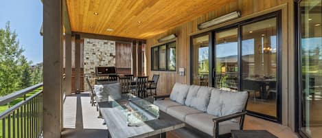 An outdoor patio with a wooden ceiling, a stone wall, a barbecue grill, and a seating area with a glass-top table and sofas.