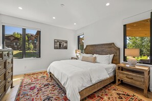 A bedroom with a large bed, a wooden headboard, a nightstand with a lamp, a dresser, and a view of a wooden cabin through a window. - Mountain Luxury Lodge w/Gondola Access 1099 (Steamboat Springs)