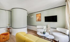Habitació d'hotel moderna amb un sofà groc, butaques blanques, una taula de centre rodona i un televisor de pantalla plana penjat a la paret.
