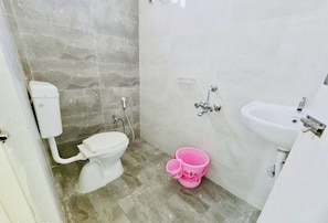 A bathroom with a toilet, a sink, and a shower. There is a pink bucket on the floor. - Deluxe Srinivasa Home Stays (Hyderabad)