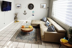 Ein Wohnzimmer mit einem beigefarbenen Sofa, einem runden Couchtisch mit Dekoration, einem gemusterten Teppich und Wanddekorationen, darunter eine Uhr und gerahmte Bilder.