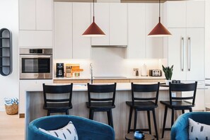 A modern kitchen with white cabinets, a built-in oven, and a bar area with four black chairs. - 2 Turns - Revelstoke Vacations (Revelstoke)